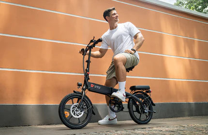 Man in white shirt and khaki shorts riding an electric bike near a brick wall.