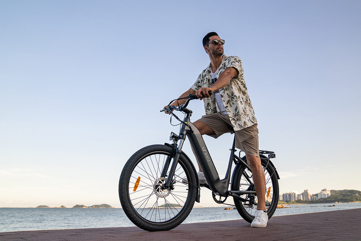 Man in stylish shirt riding the DYU C1 26 Inch City Electric Bike by the seaside.