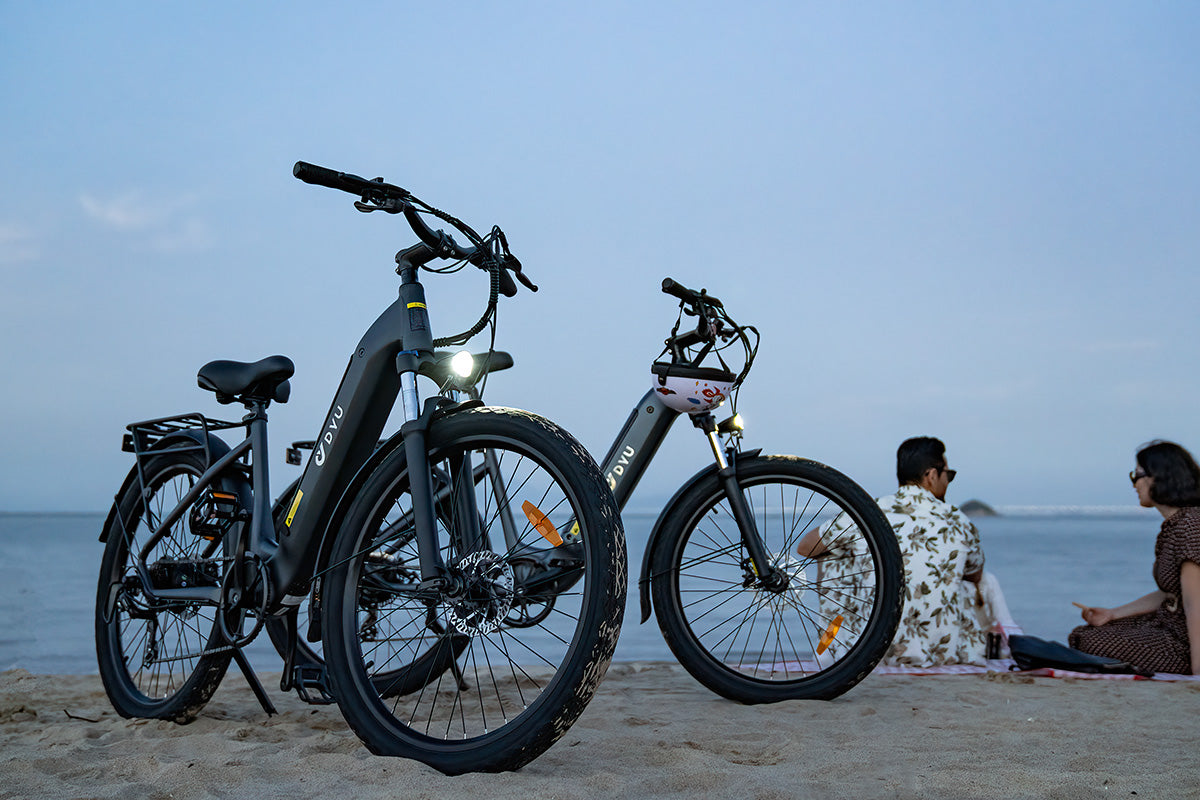 Two grey DYU C1 26 Inch City Electric Bikes on the beach with people relaxing nearby.